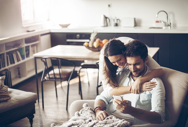 Woman standing behind man on chair hugging him and looking at his phone with him