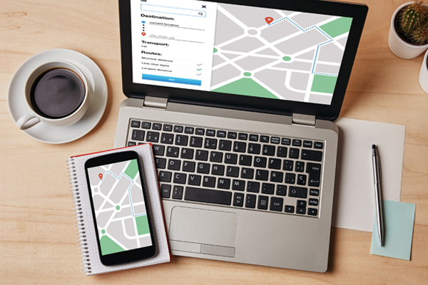 A laptop displaying a map directions on a table with a phone showing the same map and a cup of coffee, notebook, pen and paper