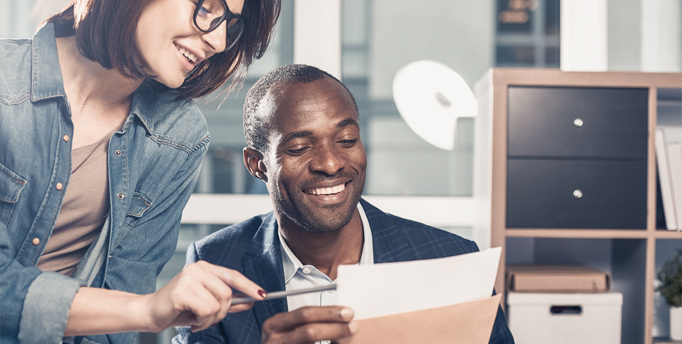 Smiling woman wearing glasses, holding a pencil, leaning over a smiling man sitting down in an office holding 2 documents