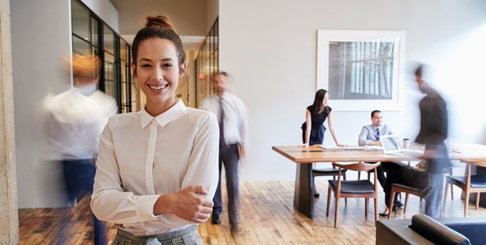 Smiling woman standing facing forward in a room with a hallway behind her and table, chairs and 5 other people in the background