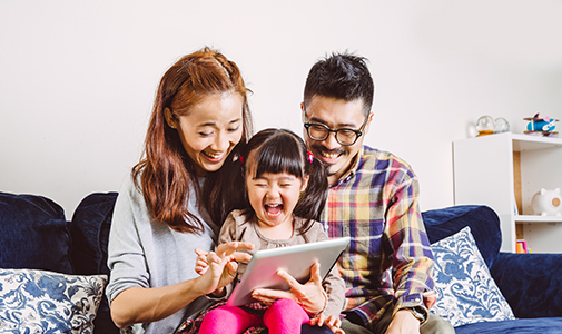 a family of three sitting on a couch looking at a tablet together 