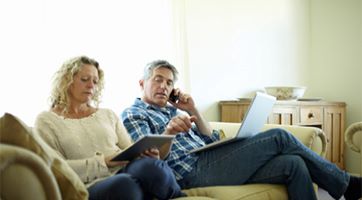 Man and woman sitting on couch, he is on the phone with a laptop of his lap, she is holding a tablet