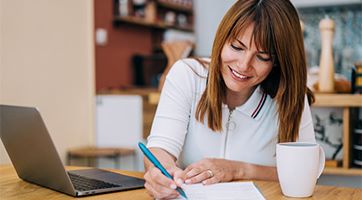 Woman sitting at able writing on a paper with a laptop and white mug sitting in front of her on the table