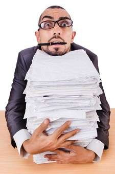 Stressed-looking man in glasses with huge stack of papers.
